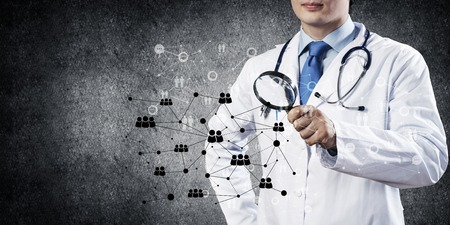 Professional and young doctor in white medical uniform looking through magnifier at social network connection while standing against dark gray wall on background.の写真素材