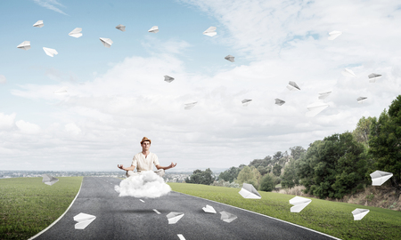 Young man keeping eyes closed and looking concentrated while meditating on cloud above the road with beautiful and breathtaking landscape on background.の写真素材