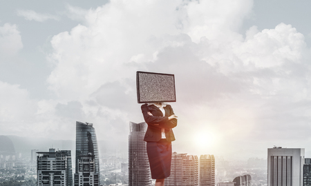 Cropped image of business woman in suit with TV instead of head keeping arms crossed while standing outdoors with cityscape view on background.の写真素材