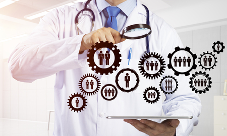 Close up of confident doctor in white sterile suit and with stethoscope looking through magnifier at team work gear structure while standing inside bright office. Medical industry conceptの写真素材