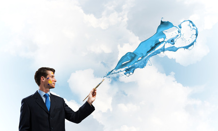 Horizontal shot of cheerful and young businessman in black suit standing with blue coloured splash against cloudy skyscape on background.の写真素材