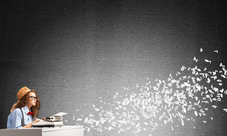 Young and beautiful woman writer in hat and eyeglasses using typing machine while sitting at the table among flying letters and against gray concrete wall on background.の写真素材