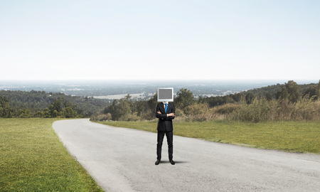 Businessman in suit with monitor instead of head keeping arms crossed while standing on the road with beautiful landscape on background.の写真素材