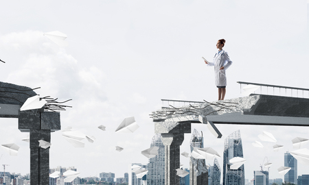 Confident medical industry employee in white uniform standing among flying paper planes at the edge of broken bridge with cityscape view on backgroundの写真素材