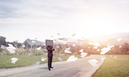 Businessman in suit with TV instead of head keeping arms crossed while standing on the road among flying paper planes with beautiful landscape on background.の写真素材