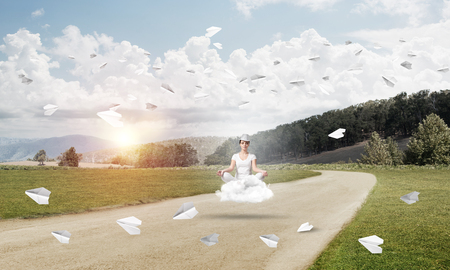 Young woman keeping eyes closed and looking concentrated while meditating on cloud above the road with beautiful and breathtaking landscape on background.の写真素材