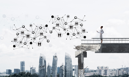 Conceptual image confident female doctor in white uniform standing at end of broken bridge and interracting with social media network structure. Medical industry concept. Cityscape on backgroundの写真素材