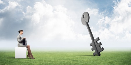 Elegant businesswoman with suitcase in hand sitting on white cube and stone key symbolの写真素材
