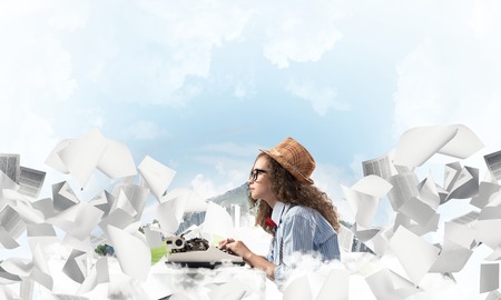 Young woman writer in hat and eyeglasses using typing machine while sitting at the table among...
