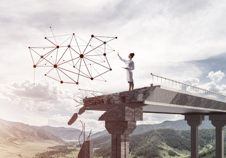 Young and professional woman doctor in white medical suit studying network structure while standing on broken bridge. Medical industry concept. Doctor using tabletの写真素材