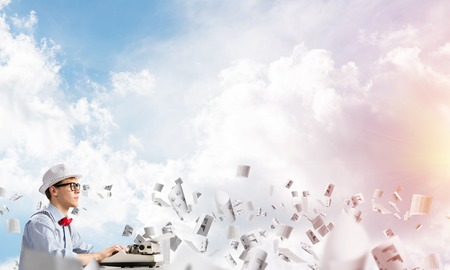 Young man writer in hat and eyeglasses using typing machine while sitting at the table among flying papers with cloudy skyscape on background.の写真素材