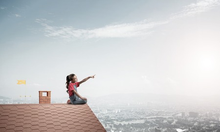 Cute kid girl sitting on house brick roof and pointing at somethingの写真素材