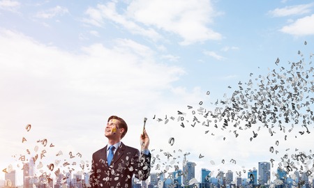 Portrait of young thoughtful man holding paintbrush in his hand and looking away while standing with flying letters and against modern cityscape view and cloudy sky on background.の写真素材