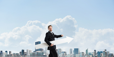 Cropped image of successful young businessman holding big white arrow and pointing away while standing against cityscape view on backgroundの写真素材