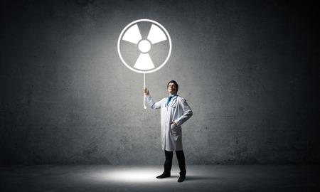 Young medical industry employee standing inside empty room and interracting with glowing radioactive symbol with gray wall on background.の写真素材