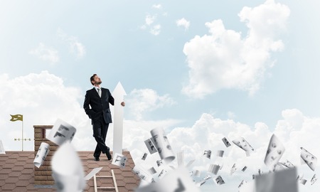 Conceptual image of young businessman in suit looking upside and holding big white arrow in hand while standing on brick roof among flying papers and cloudy skyscape view on background.の写真素材