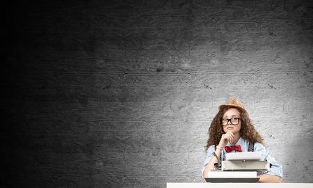 Young and beautiful woman writer in hat and eyeglasses using typing machine while sitting at the table against gray concrete wall on background.の写真素材