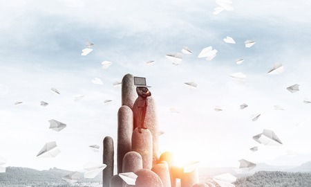 Businessman in suit with laptop instead of head keeping arms crossed while standing on the top of stone column among flying paper planes with beautiful skyscape on background. 3D rendering.の写真素材
