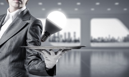 Cropped image of waitress's hand in white glove presenting glowing lightbulb on metal tray as symbol of new idea with office view on background.の写真素材