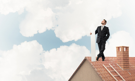Conceptual image of young businessman in suit looking upside and holding big white arrow in hand while standing on brick roof with cloudy skyscape view on background.の写真素材