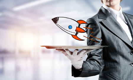 Cropped image of waitresss hand in white glove presenting sketched flying missile on metal tray with office view on background.の写真素材