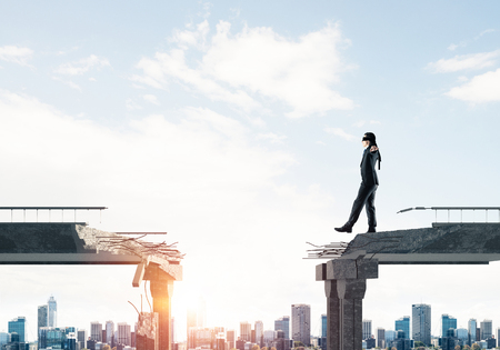 Businessman walking blindfolded on concrete bridge with huge gap as symbol of hidden threats and risks. Cityscape view with sunlight on background. 3D rendering.の写真素材