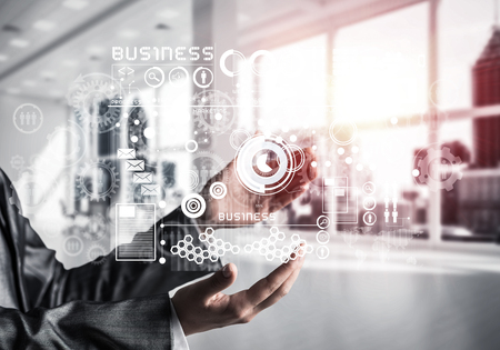 Cropped image of business woman in suit with business interface and media icons in her hands. Sunlight and office view on background. Mixed media.の写真素材