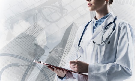 Portrait of young female doctor in white sterile suit holding notebook in hands while standing against modern city view and medical equipment on background. Double exposureの写真素材