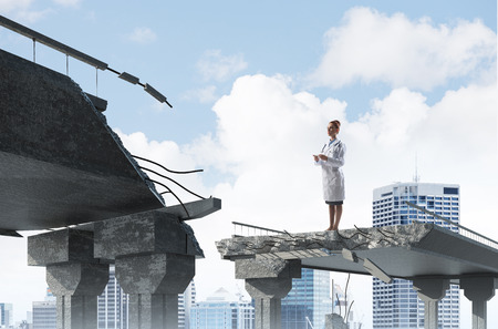 Horizontal shot of young but professional female doctor holding tablet in hands while standing at the end of broken bridge with cityscape view on background. Medical industry conceptの写真素材