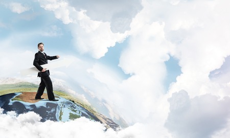 Young successful businessman in suit keeping big white arrow and pointing away while standing on Earth glove and cloudy sky on background.の写真素材