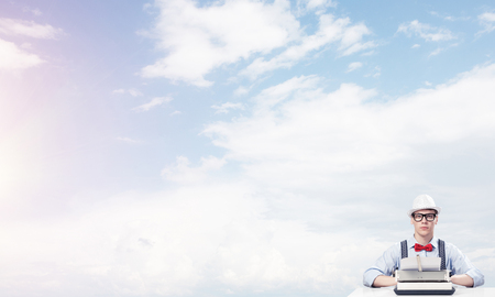 Young man writer in hat and eyeglasses using typing machine while sitting at the table with cloudy skyscape on background.の写真素材