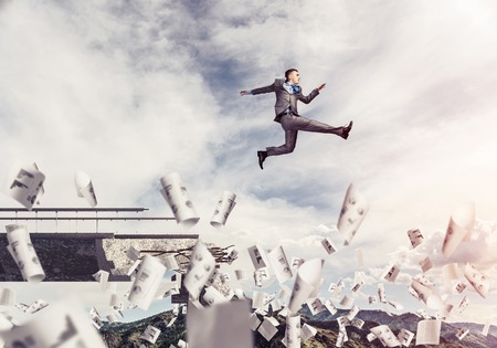 Businessman jumping over gap in bridge among flying papers as symbol of overcoming challenges. Skyscape with sunlight and nature view on background. 3D rendering.の写真素材