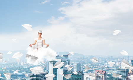 Woman in white clothing keeping eyes closed and looking concentrated while meditating on cloud among flying paper planes with cityscape view on background.の写真素材