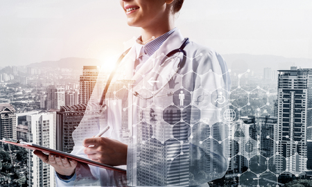 Double exposure of cheerful woman doctor standing with pen and notebook in hands and modern cityscape view and medical interface icons on background. Medical industy conceptの写真素材