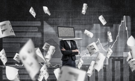 Business woman in suit with laptop instead of head keeping arms crossed while standing against flying papers and analytical charts drawn on dark wall on background.の写真素材
