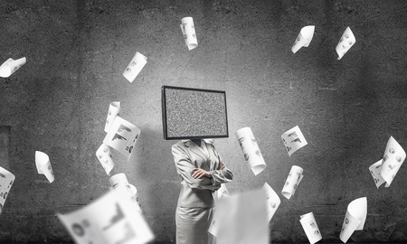 Business woman in suit with TV instead of head keeping arms crossed while standing among flying papers and with gray dark wall on background.の写真素材
