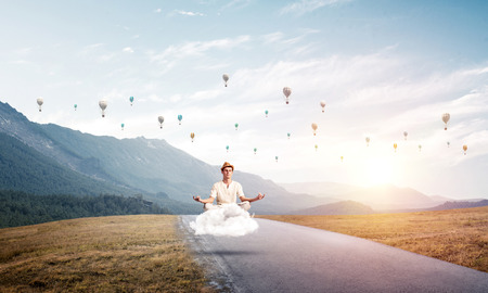 Young man keeping eyes closed and looking concentrated while meditating on cloud above the road with beautiful and breathtaking landscape on background.の写真素材