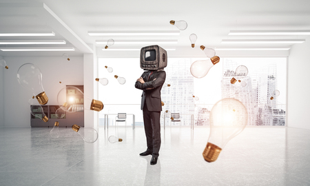 Businessman in suit with an old TV instead of head keeping arms crossed while standing among flying and glowing lightbulb inside office building. 3D rendering.の写真素材
