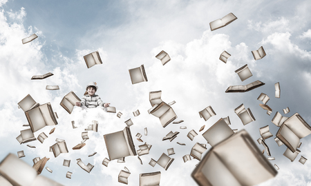 Young little boy keeping eyes closed and looking concentrated while meditating among flying books in the air with cloudy skyscape on background.の写真素材