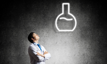 Young confident doctor in white medical uniform interracting with glowing vial symbol whie standing against dark gray wall on background.の写真素材