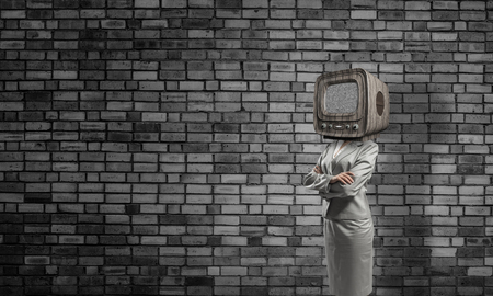 Business woman in suit with old TV instead of head keeping arms crossed while standing inside empty room with gray dark wall on background.の写真素材