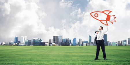 Confident and young businessman in suit starting launching a big rocket from his hand while standing on green meadow and city view on background.の写真素材