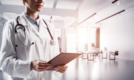 Close up of successful female doctor standing with tablet in hands indoors of white medical office building. Young woman doctor. Medical industry conceptの写真素材