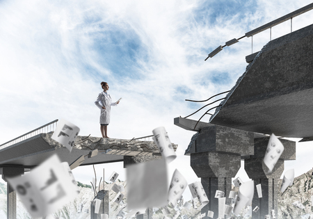 Confident female doctor using tablet while standing among flying papers at the edge of broken bridge with landscape view on background. Medical industry conceptの写真素材
