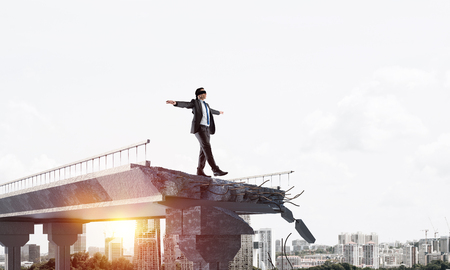 Businessman walking blindfolded on concrete bridge with huge gap as symbol of hidden threats and risks. Cityscape and sunlight on background. 3D rendering.の写真素材