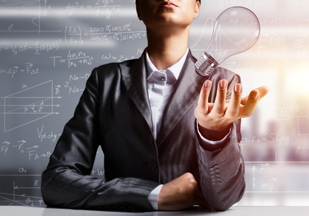 Businessman in suit presenting lightbulb in hand as symbol of new idea, office view on background. 3D rendering.の写真素材