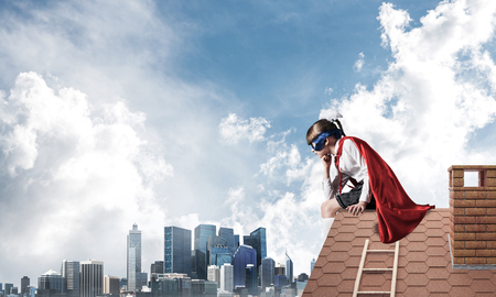 Little sad child in mask and cape sitting on roof edge and looking down. Mixed mediaの写真素材