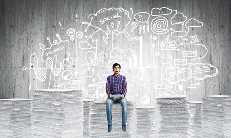 Young man in casual wear sitting on pile of documents with business-analytical information on background. Mixed media.の写真素材