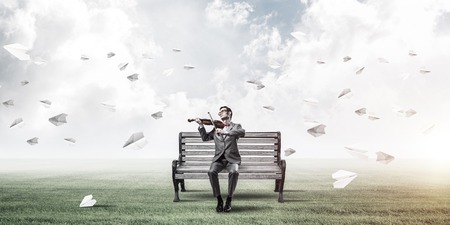 Young man wearing suit and glasses sitting on bench and playing violinの写真素材
