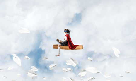 Little child in mask and cape floating on book in sky and readingの写真素材